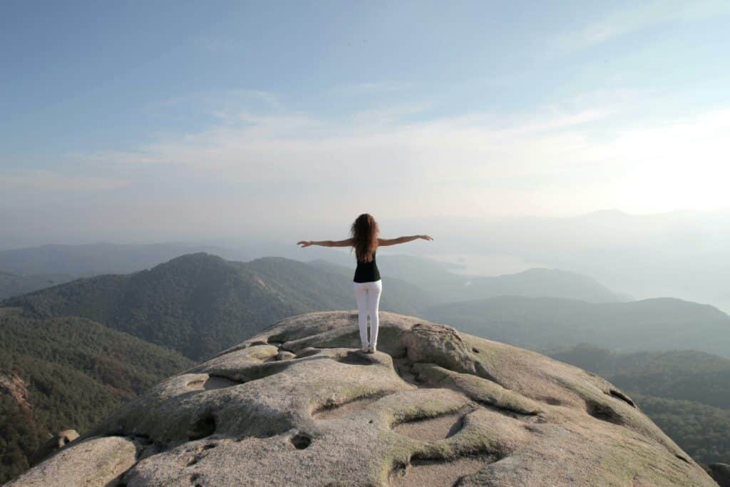 A woman stands and embraces freedom on a mountain peak overlooking a vast landscape.